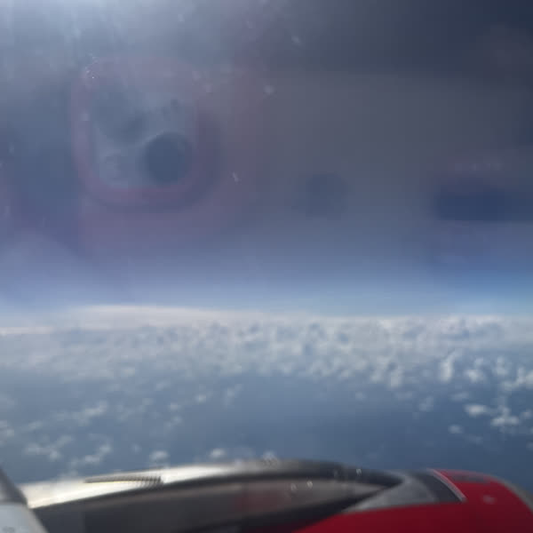 Clouds and a red airplane wing through a scratched cabin window